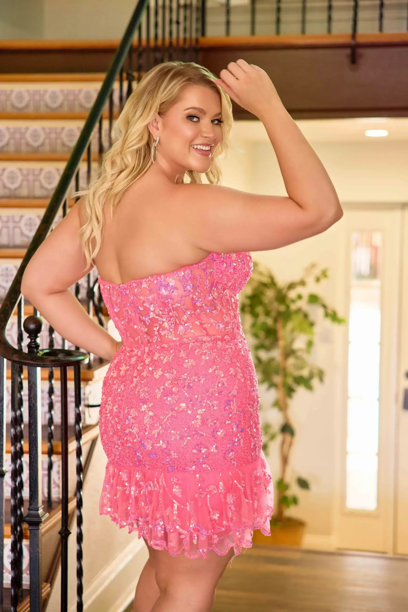 A woman wearing a strapless pink dress adorned with floral embellishments stands at the bottom of a staircase, smiling and posing with her hand on her hip.