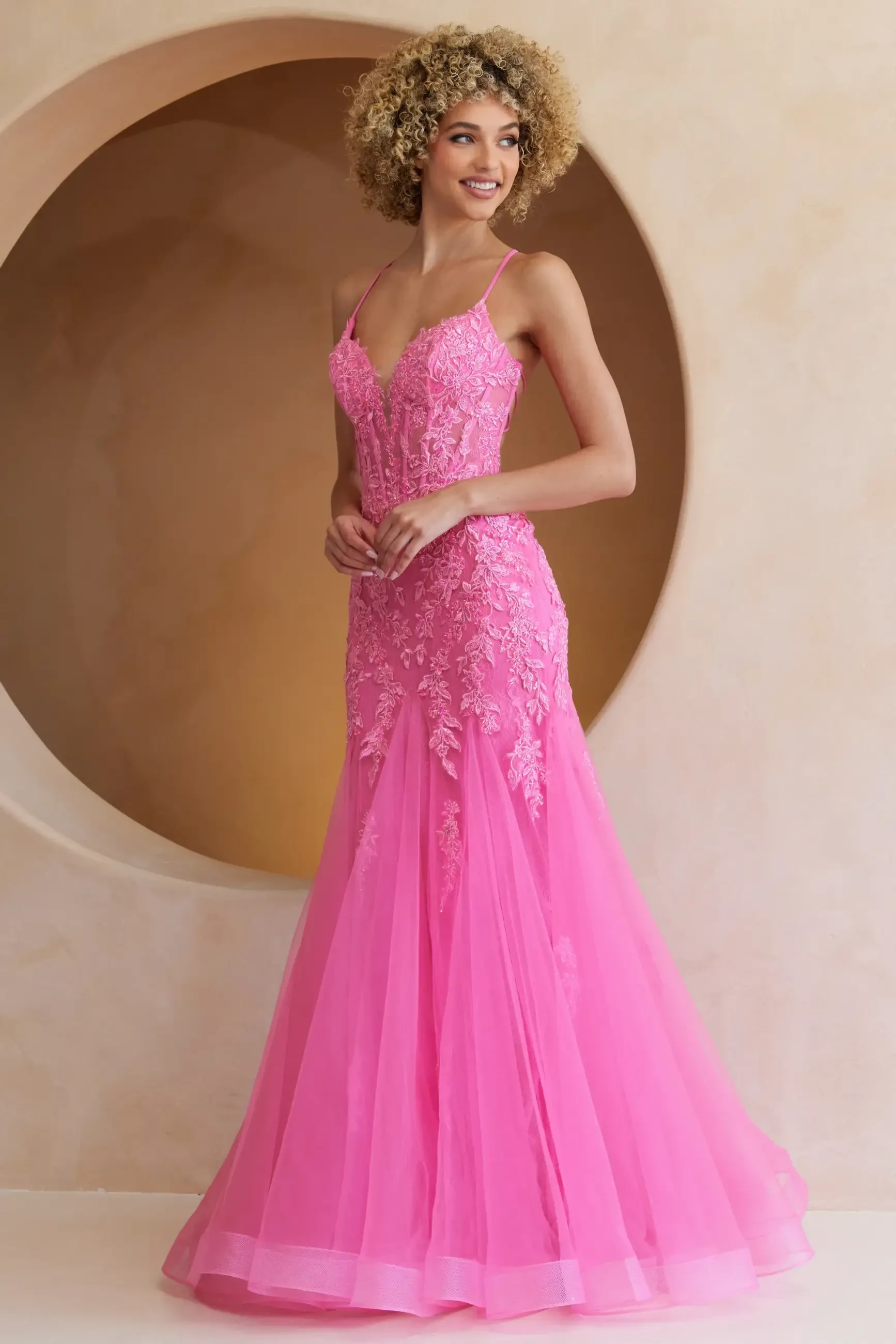 A woman in a vibrant pink lace gown stands gracefully in front of a beige circular backdrop. She's smiling softly, exuding elegance and poise.