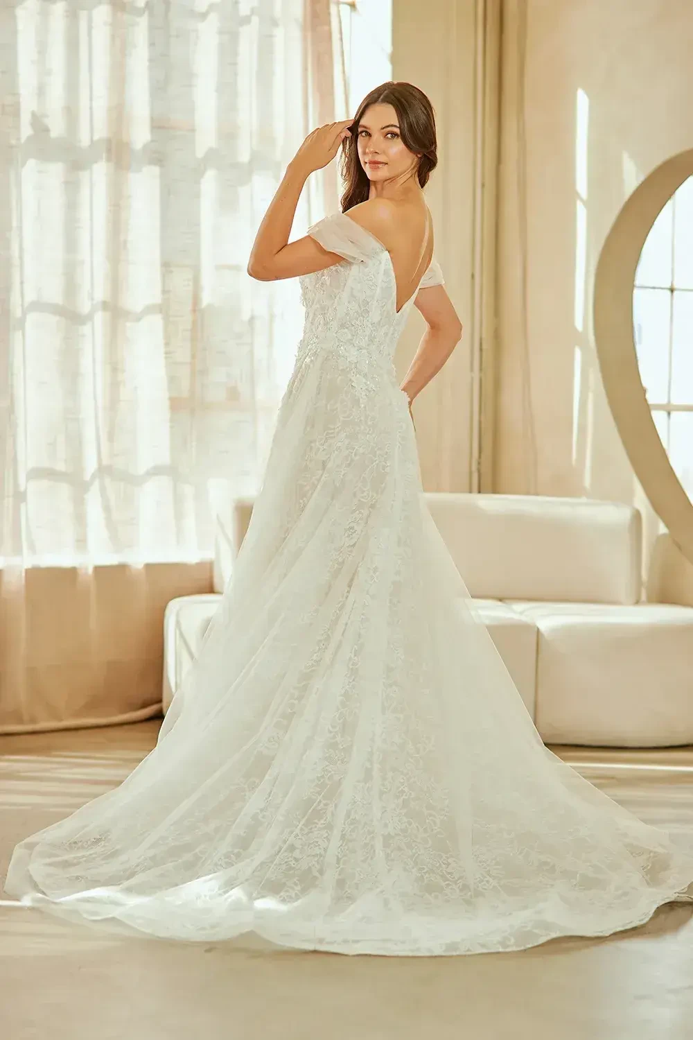 A woman in a white off-the-shoulder wedding dress is standing with her back to the camera, showcasing the gown's elegant design. The setting is bright, with soft natural light filtering through sheer curtains.