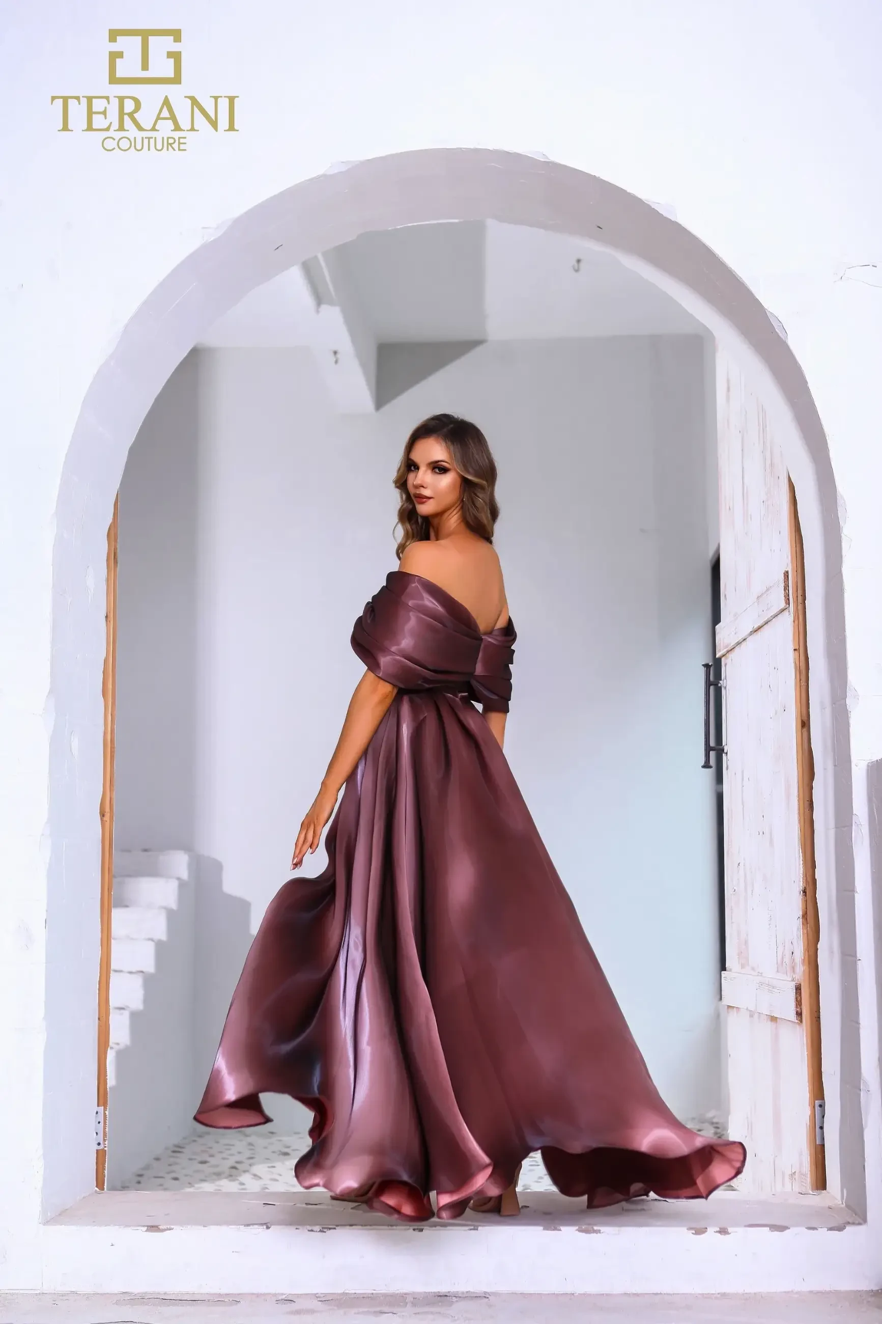 Model is wearing an espresso A-line liquid organza gown with a structured off-the-shoulder neckline and a thigh-high front slit.