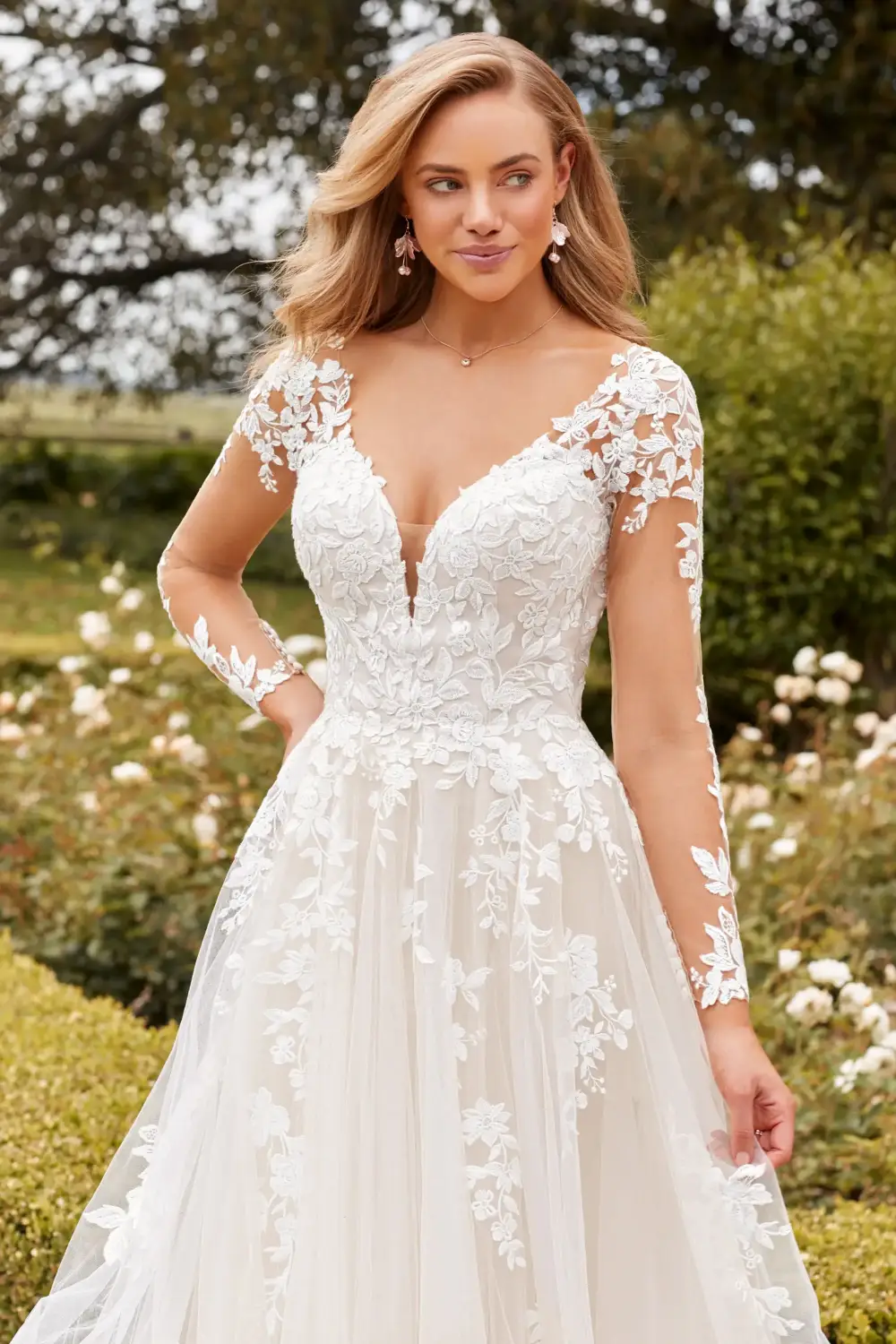 A woman wearing a beautiful wedding dress with lace details and a plunging neckline, standing outdoors in a garden.