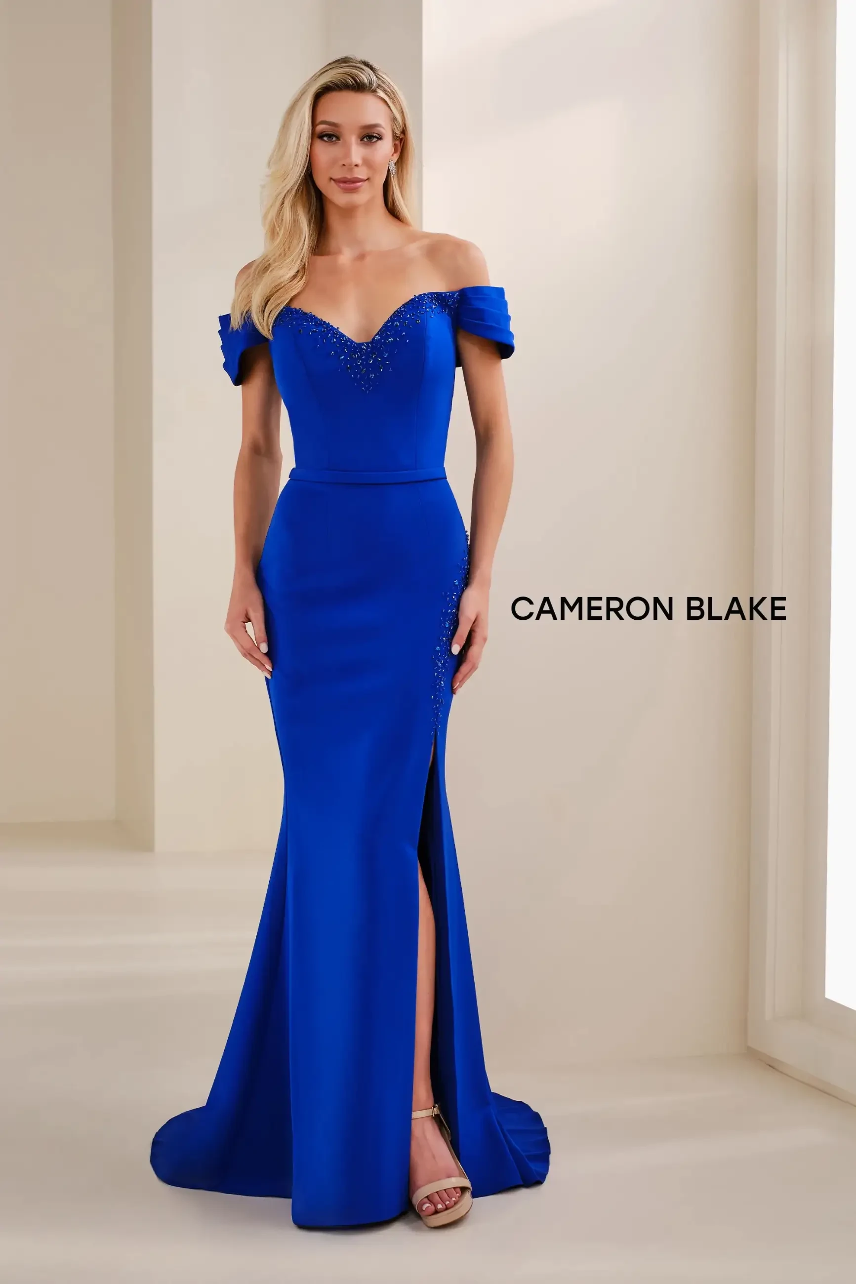 Model is wearing a royal blue crepe mermaid gown with a sweetheart neckline, delicate beading, and off-the-shoulder sleeves.