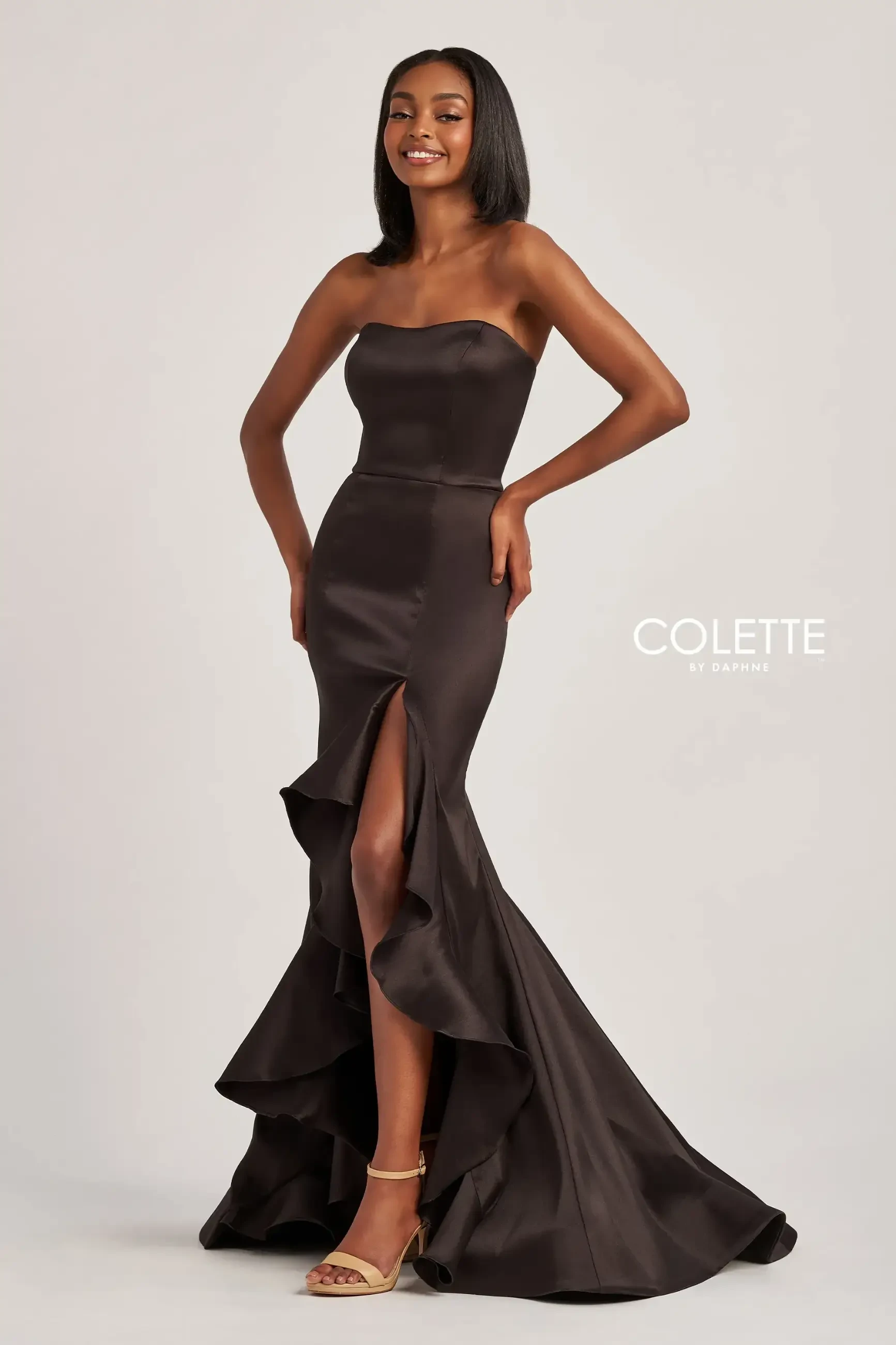 A woman wearing an elegant strapless chocolate brown gown with a high slit and ruffled train. She smiles, conveying confidence and grace.