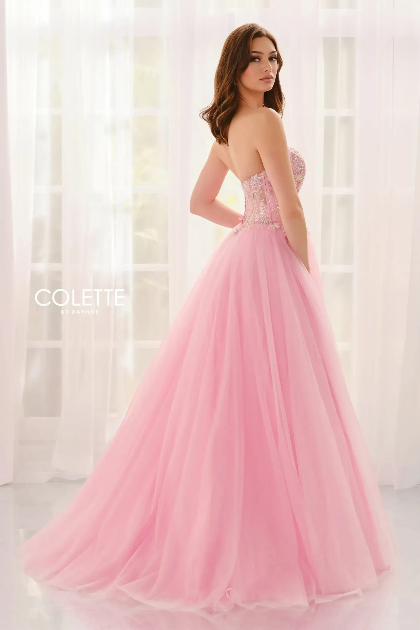 A woman in a pink strapless ball gown with a beaded bodice stands gracefully in front of sheer white curtains, exuding elegance and poise.