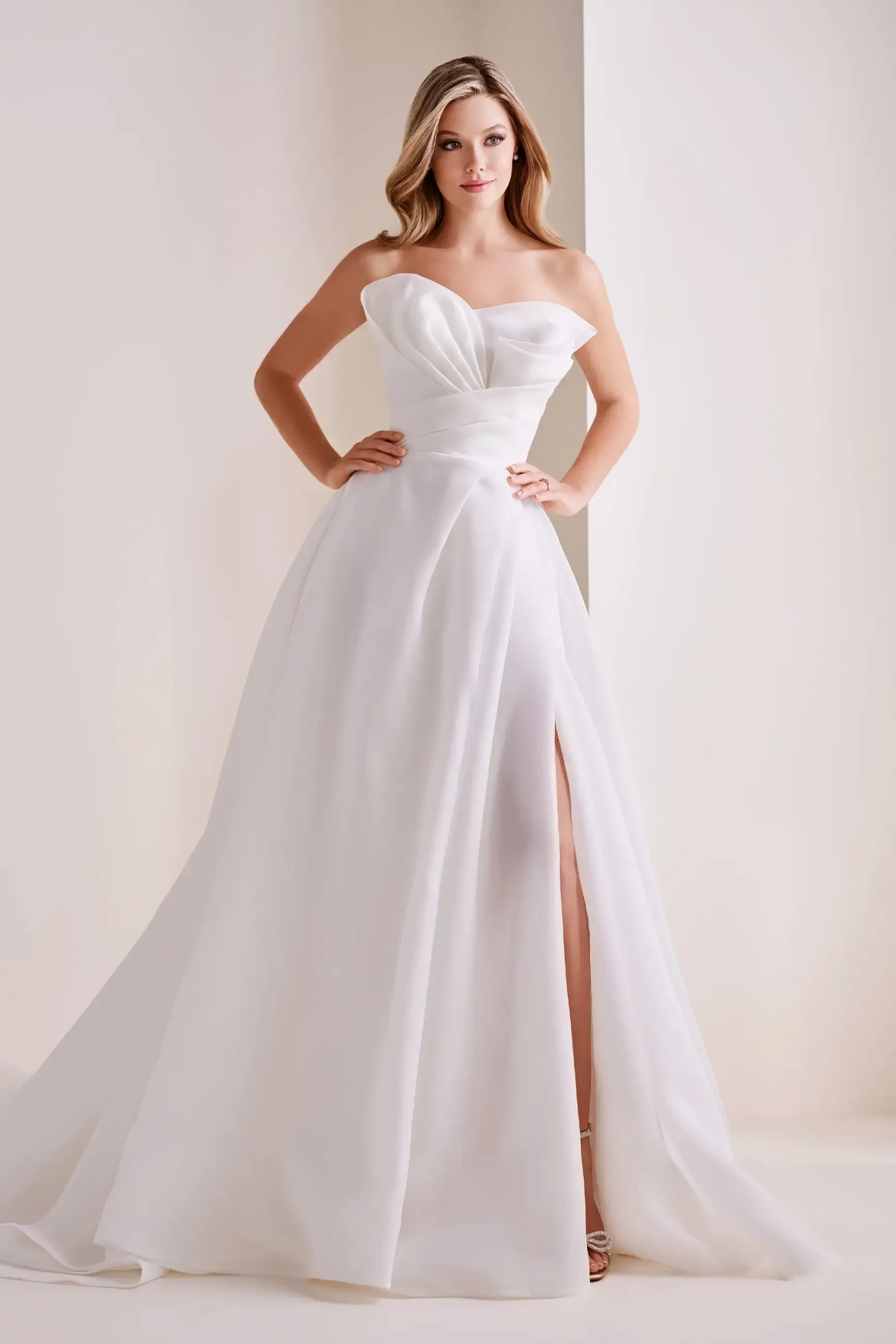 A woman in an elegant, strapless white gown with a fitted bodice and flowing skirt stands confidently. The dress has a high slit, exuding sophistication.