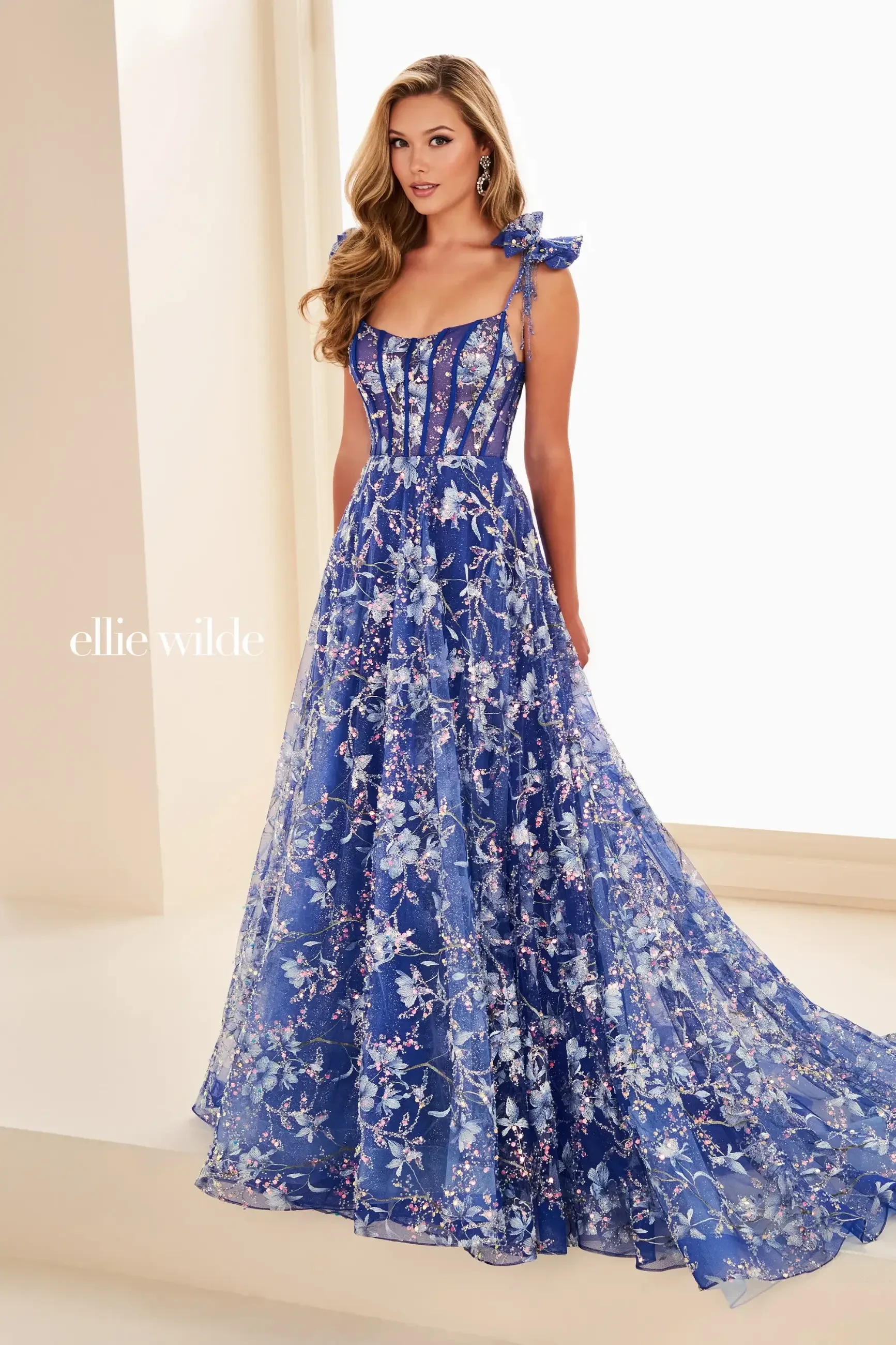 A woman in a flowing blue floral gown stands elegantly in a bright room. The dress features intricate embroidery and shoulder bows, creating a sophisticated and graceful look.