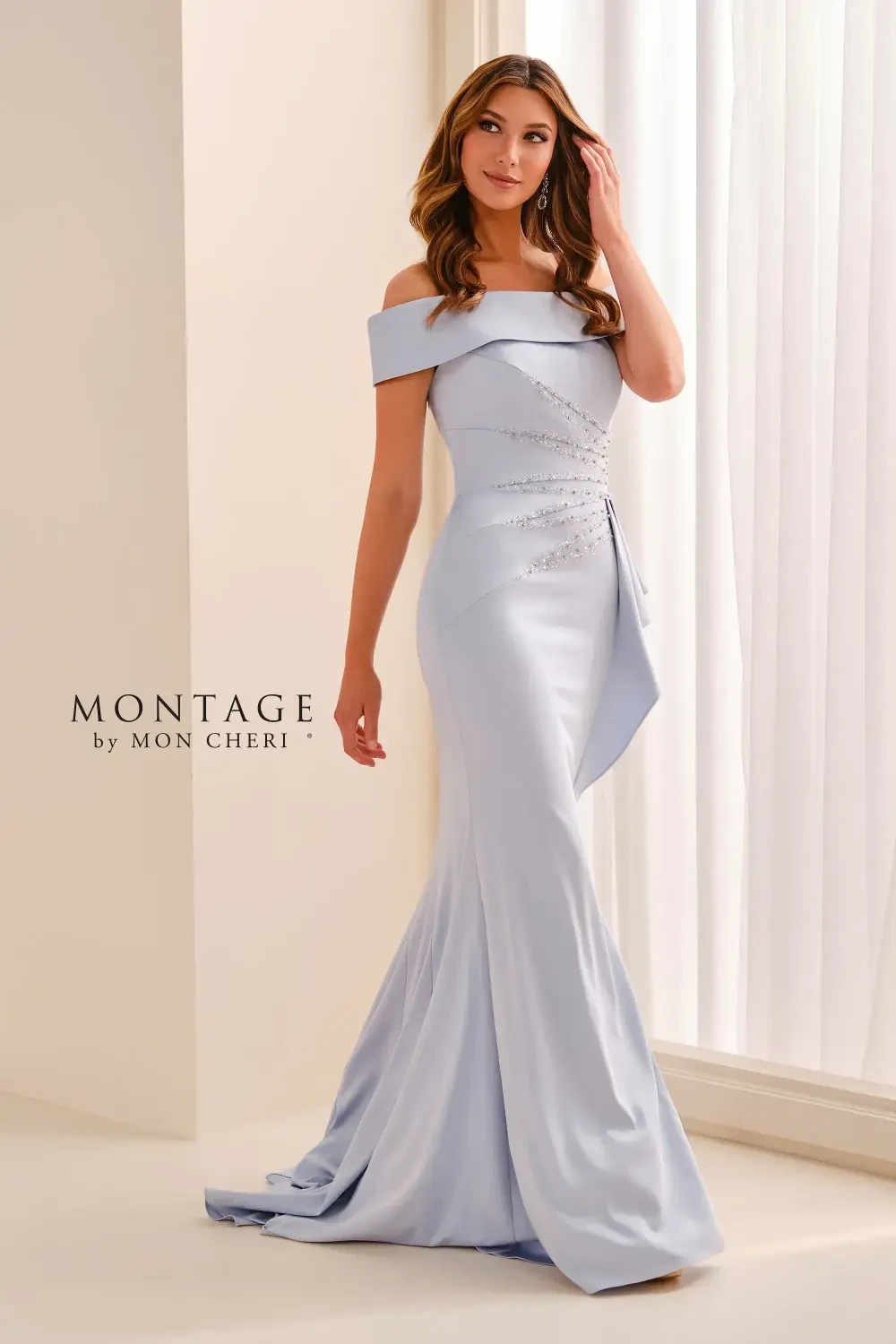 A model wearing a light blue off-the-shoulder evening gown with elegant draping and beaded detailing, standing gracefully in a well-lit room.