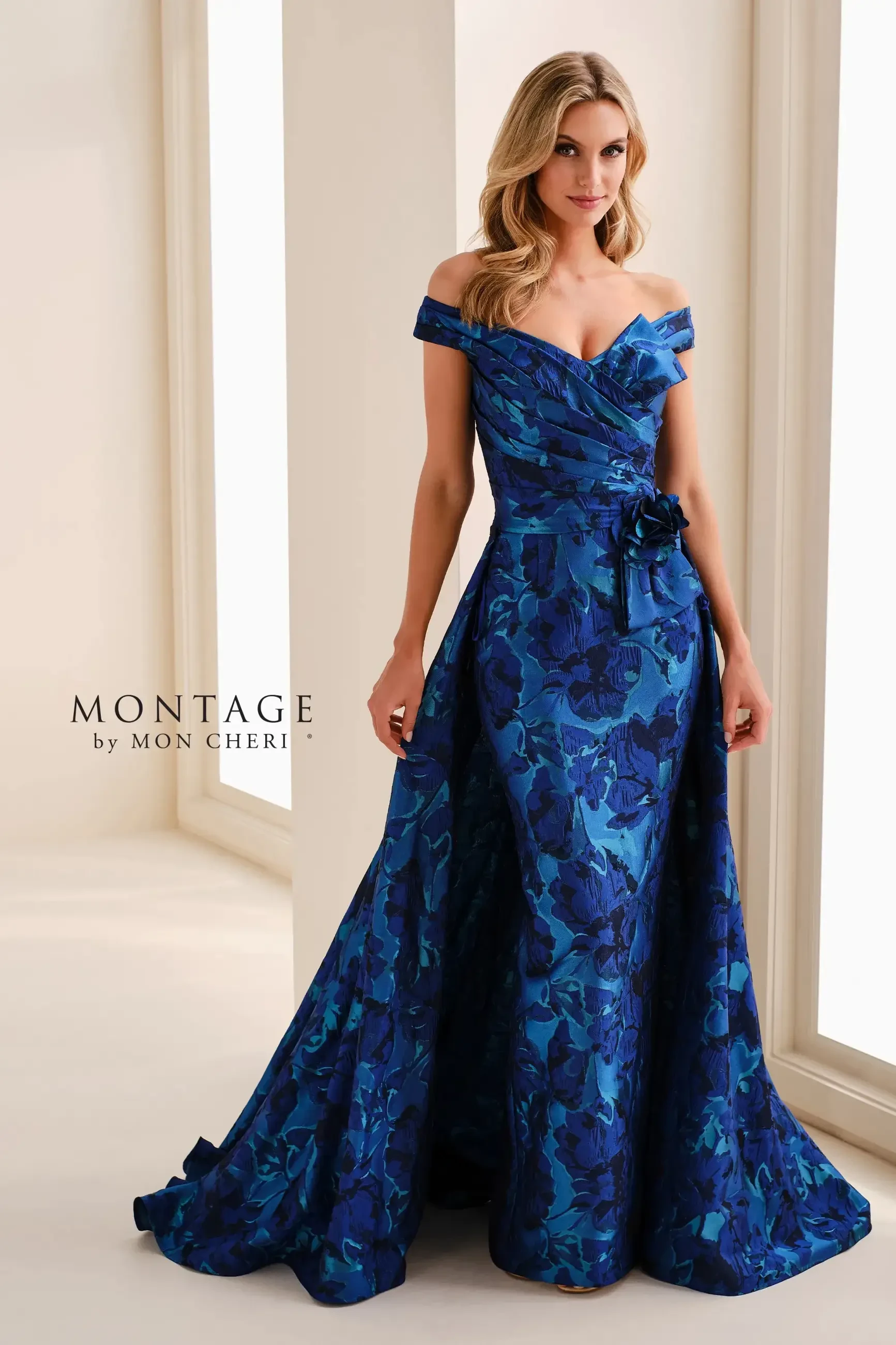 A woman wearing a formal, off-the-shoulder blue gown with floral patterns, standing elegantly in a well-lit setting.