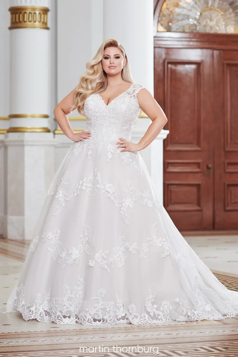 A model wearing a romantic, voluminous white wedding gown with lace detailing, posing in an elegant setting.