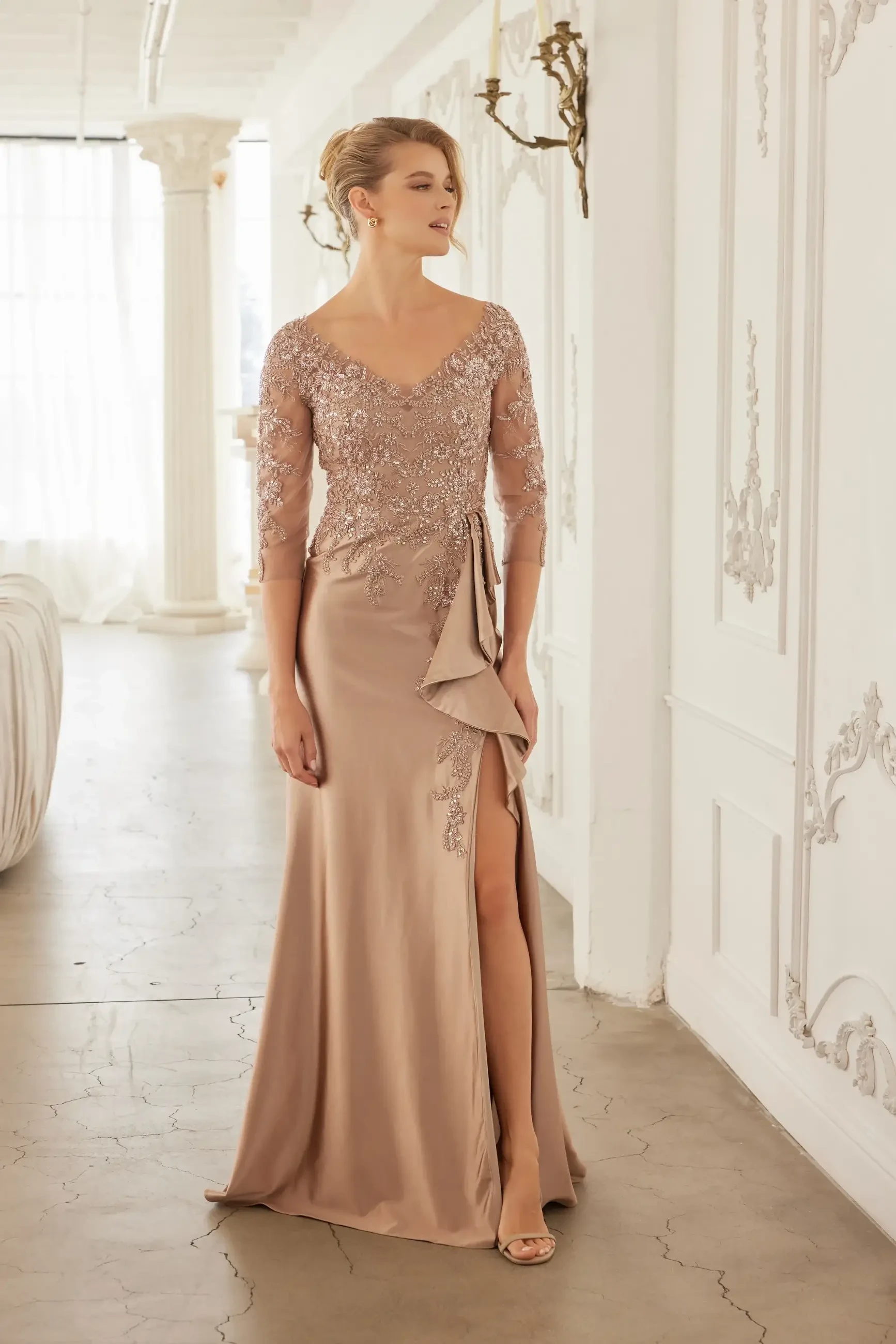 A model wearing an elegant, formal gown with a long skirt and intricate embellishments, standing in a stylish indoor setting. The dress has a deep V-neckline and features lace sleeves.