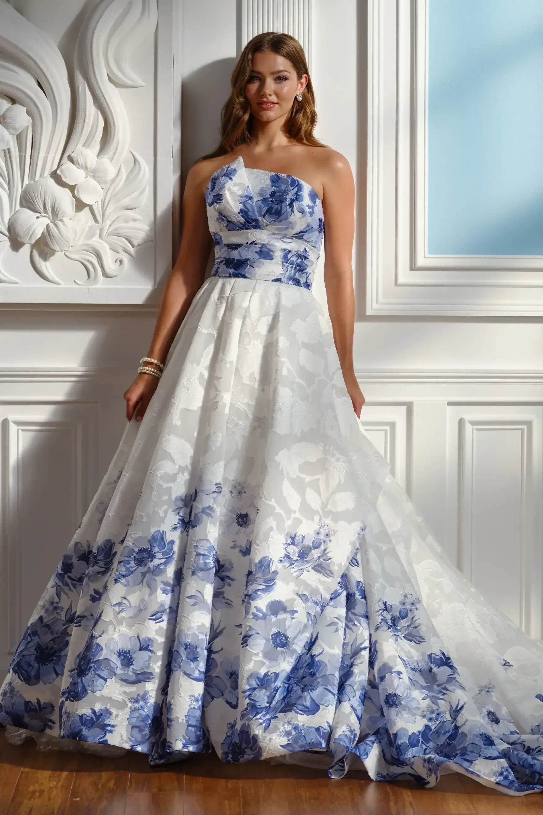 Woman in elegant, strapless white gown with blue floral patterns, standing against a white textured wall and soft blue window. The mood is serene and sophisticated.