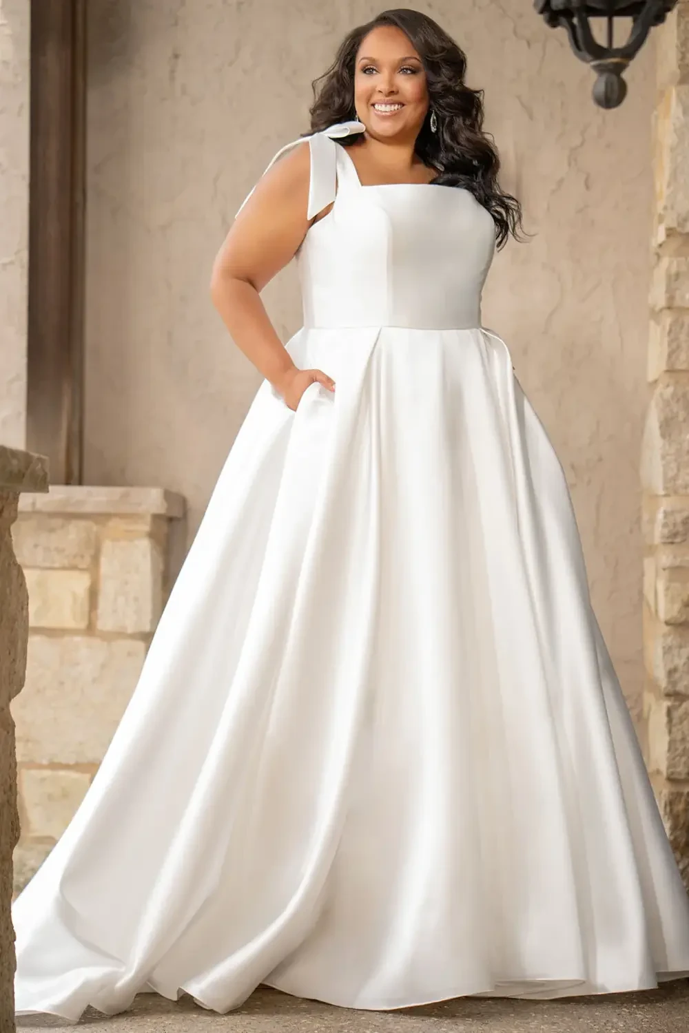A woman in a white wedding dress with a square neckline and pockets, standing outdoors and smiling.