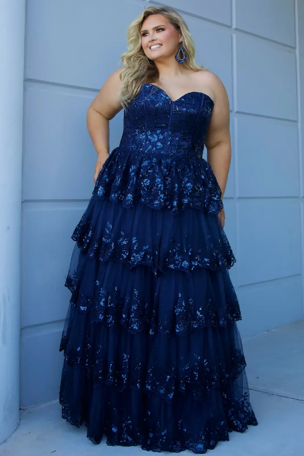 Woman in a strapless navy blue gown with sequin details and layered tulle stands against a light wall. She is smiling, exuding elegance and confidence.
