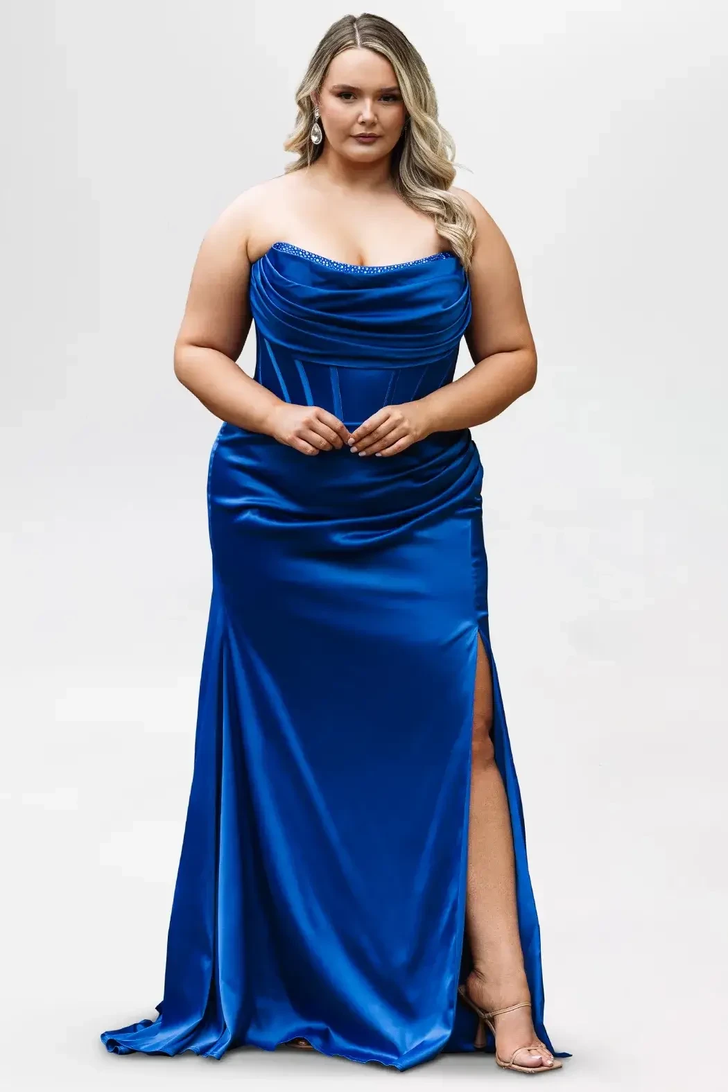 Woman in a strapless, royal blue satin gown poses against a plain background. The dress has a high slit, and she exudes confidence and elegance.