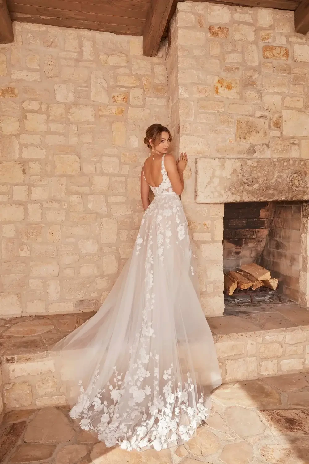 A woman in a beautiful, floral wedding dress stands gracefully with her back to the camera, positioned near a stone wall and fireplace.
