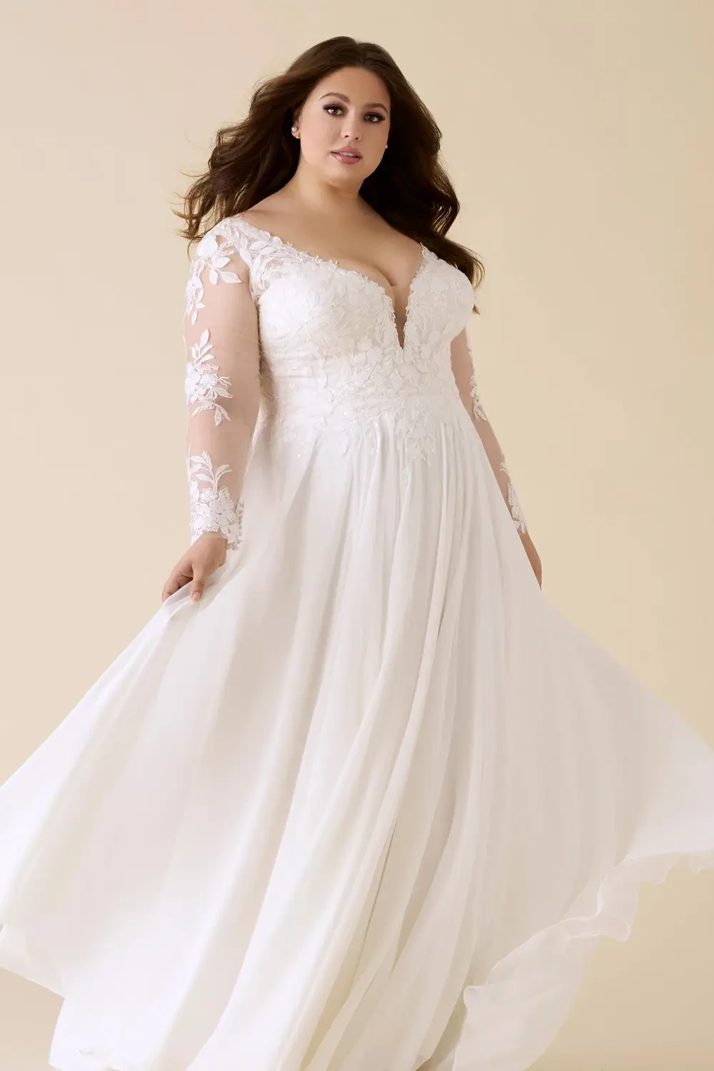 A woman wearing a white bridal gown with long sleeves, featuring floral lace details and a plunging neckline, twirling gracefully against a light beige background.