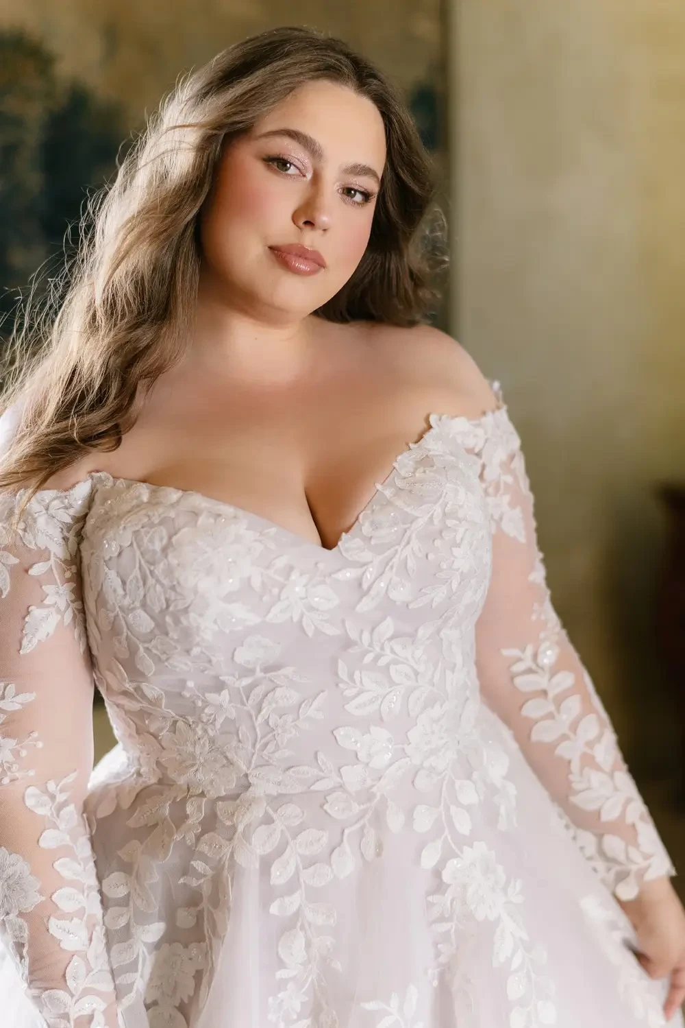 A woman with long, wavy hair wearing an off-the-shoulder wedding dress with floral lace details. She is gazing directly at the camera.