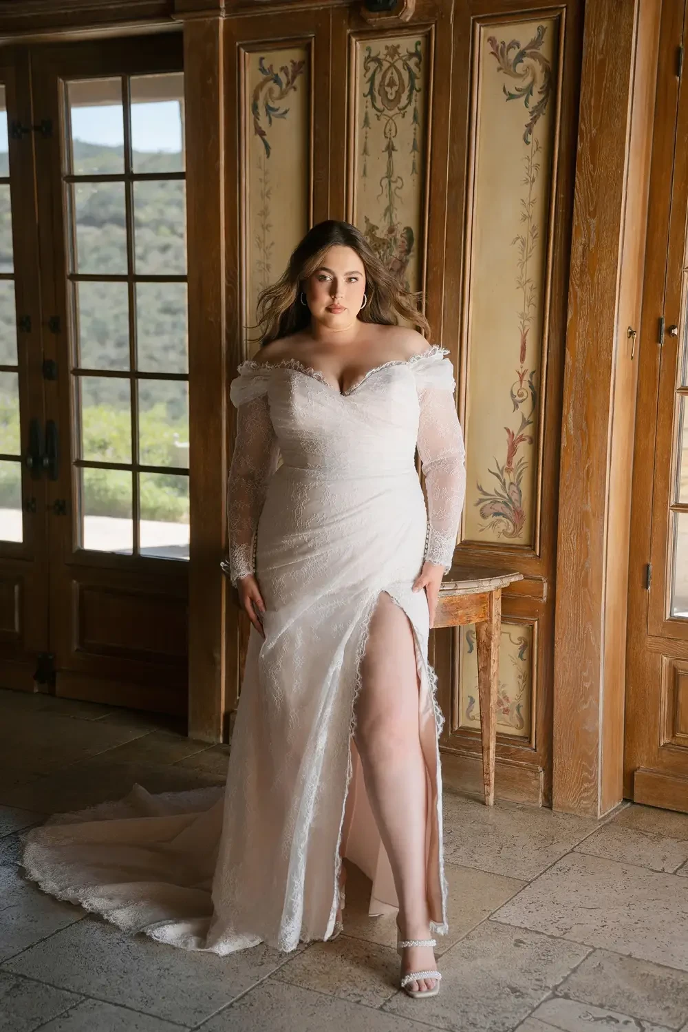 A plus-size bride in a white wedding dress with a thigh-high slit, standing indoors near large windows with a view of greenery.